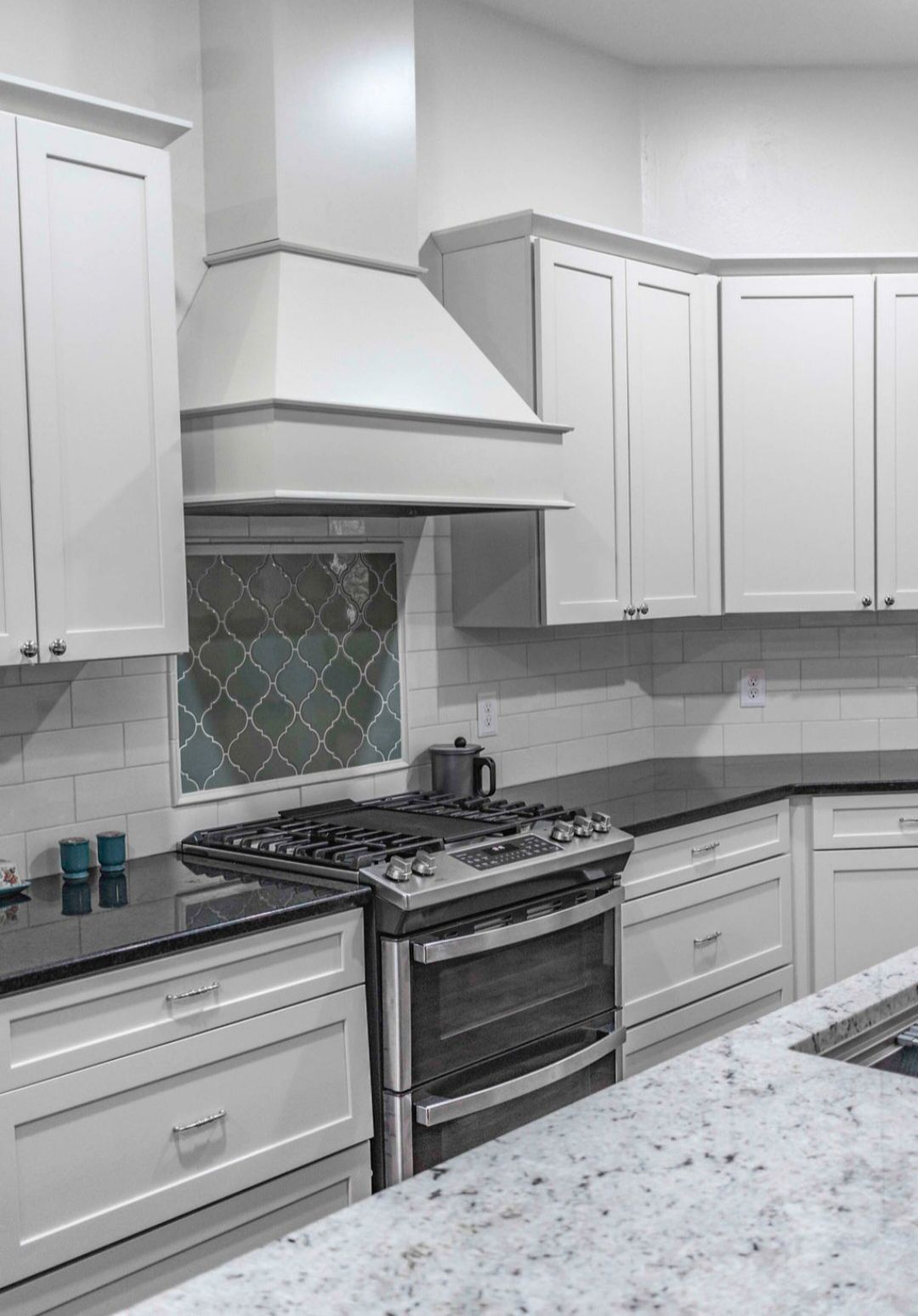 White kitchen with stove, range hood, cabinets, black countertop, and a diamond-patterned window.