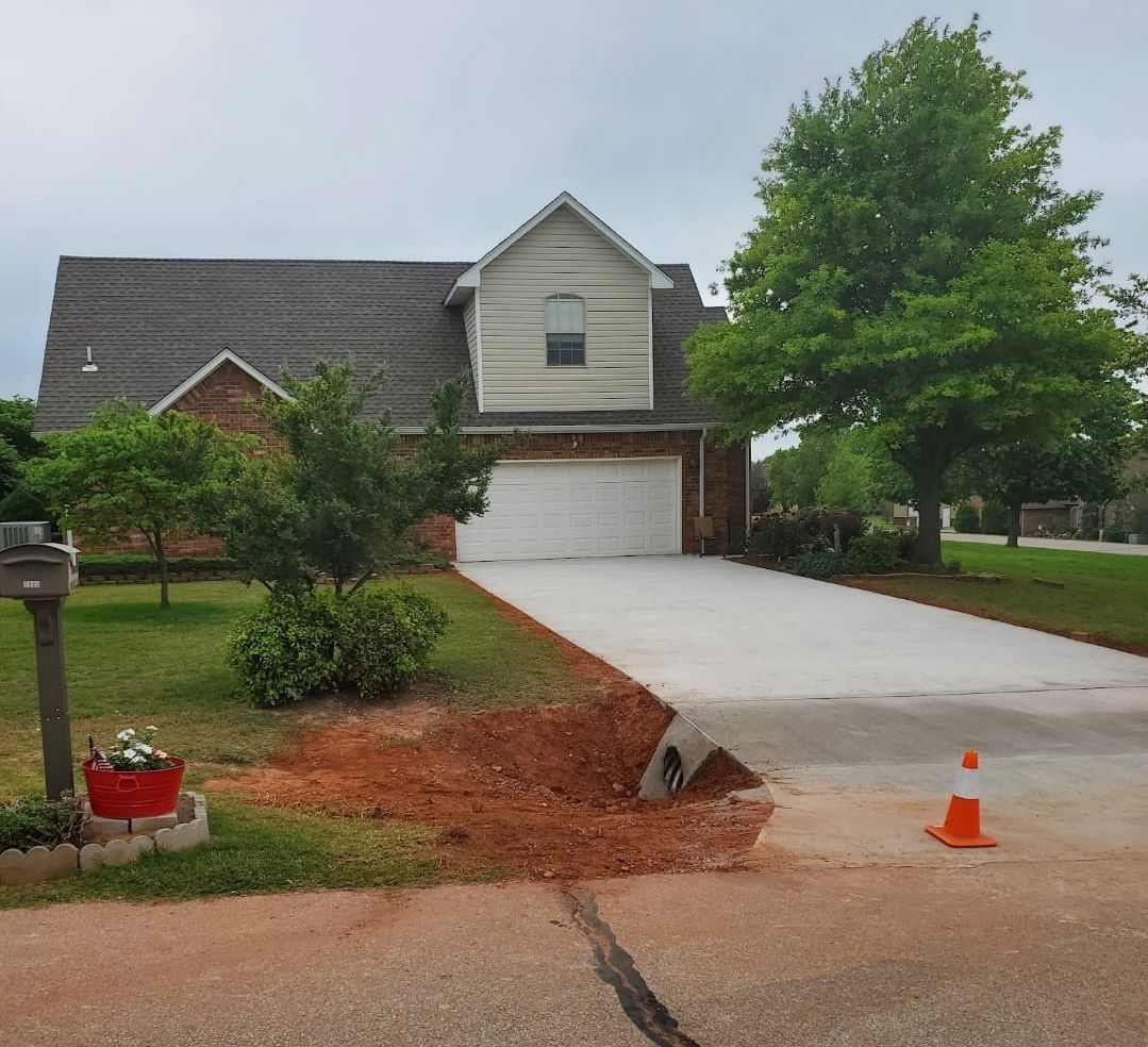 A house with a driveway that is damaged. Orange cone, dirt, and red dirt are visible.