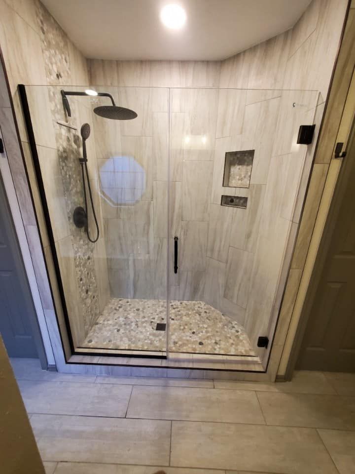 Modern shower with glass doors, black fixtures, pebble floor, and light beige tile walls.