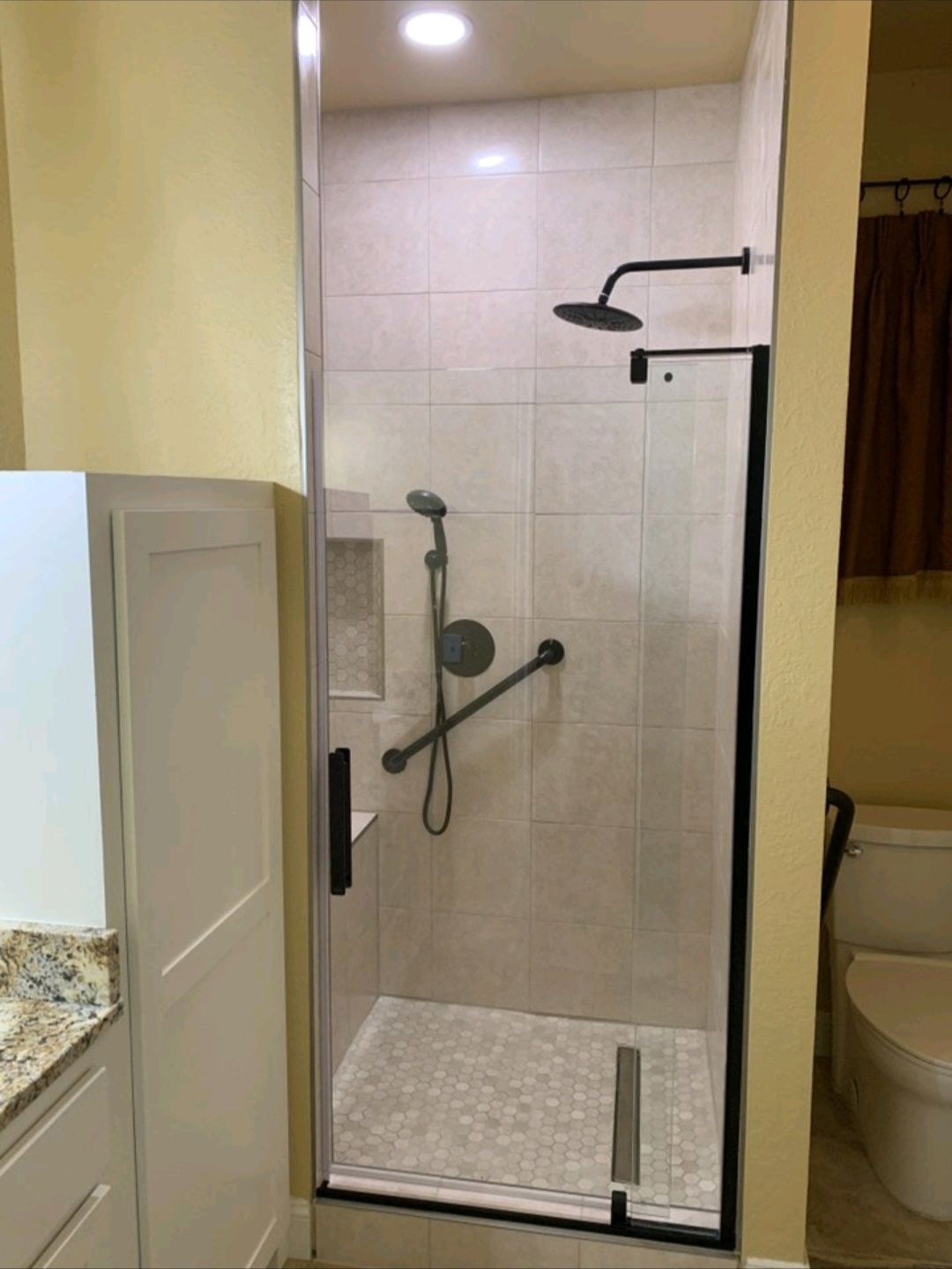 Accessible shower with grab bar, handheld shower, and pebble floor. Black fixtures on beige tiled walls.