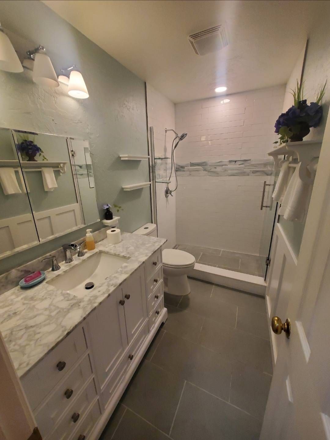Bathroom with white vanity, toilet, and shower. Pale blue walls, gray tile floor, and white trim.