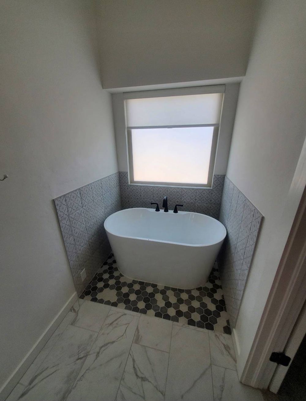 Freestanding bathtub in a bathroom niche with patterned floor and small window.