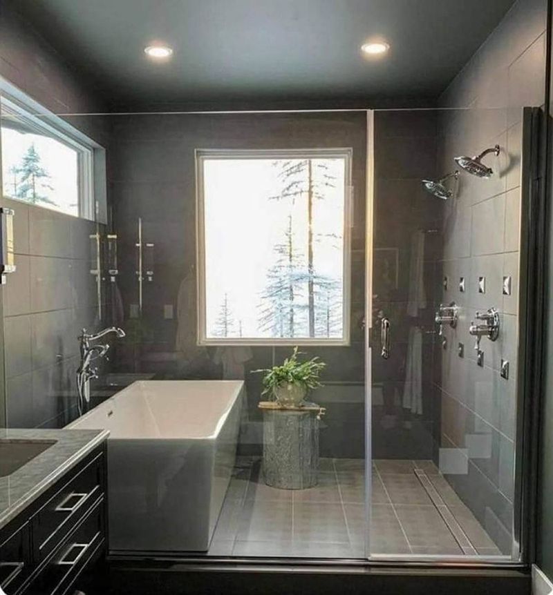 Modern gray bathroom with a glass shower, soaking tub, window with a snowy view, and dark cabinets.