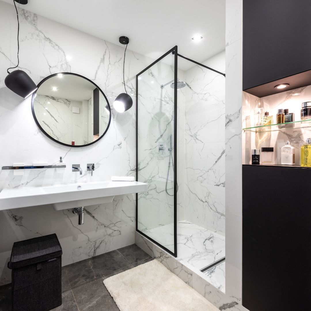 Modern bathroom with marble-look walls, glass shower, floating sink, black accents, and round mirror.