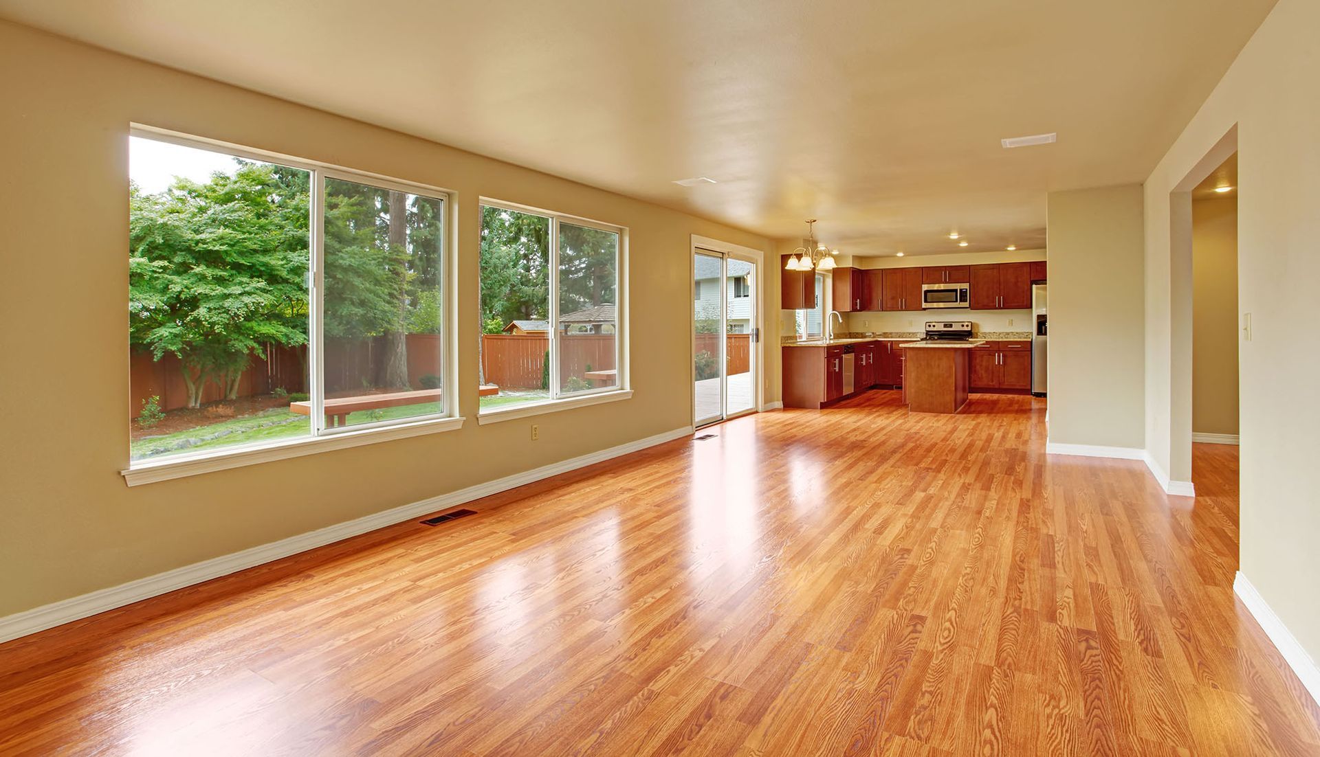 Spacious living room with hardwood floors, windows looking onto a yard, and view of a kitchen.