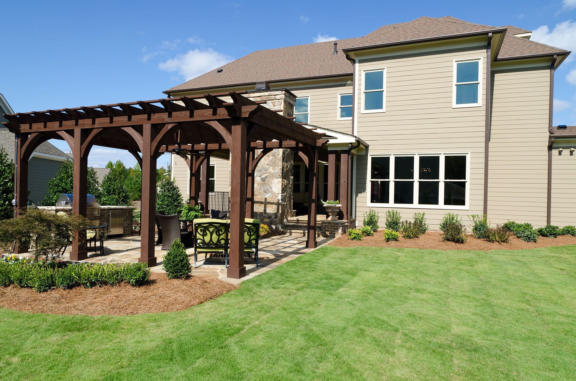 Brown pergola over a patio with outdoor furniture next to a two-story beige house on a sunny day.