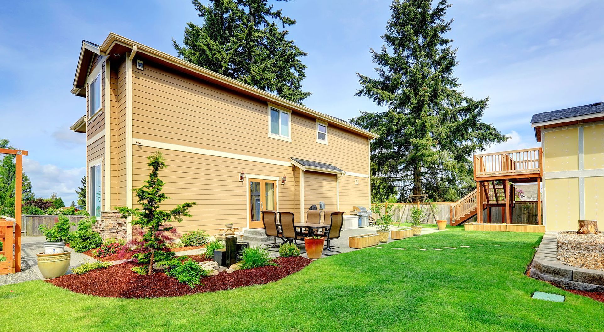 Two-story tan house with backyard patio furniture and a small pond, lush green lawn, and a blue sky.