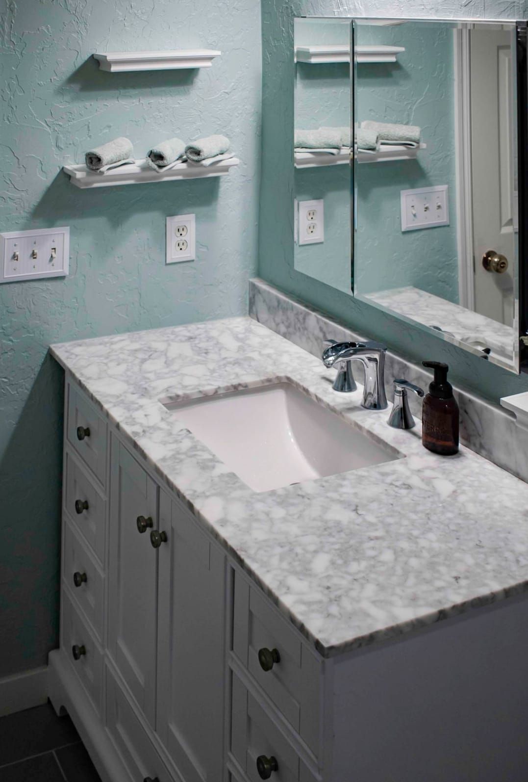 White bathroom vanity with marble countertop and sink, white towels on shelves, mirror. Teal walls.