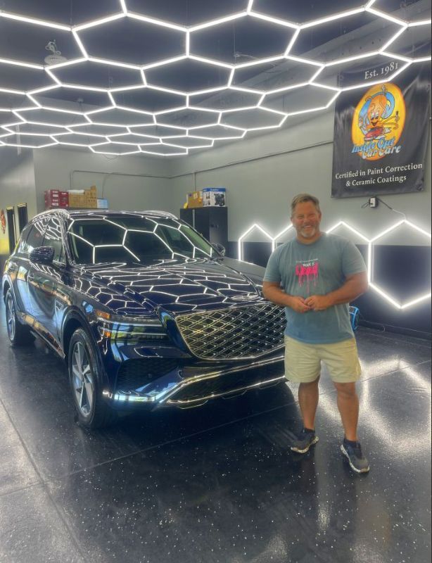 Man stands with a shiny blue SUV in a well-lit garage with honeycomb ceiling lights.