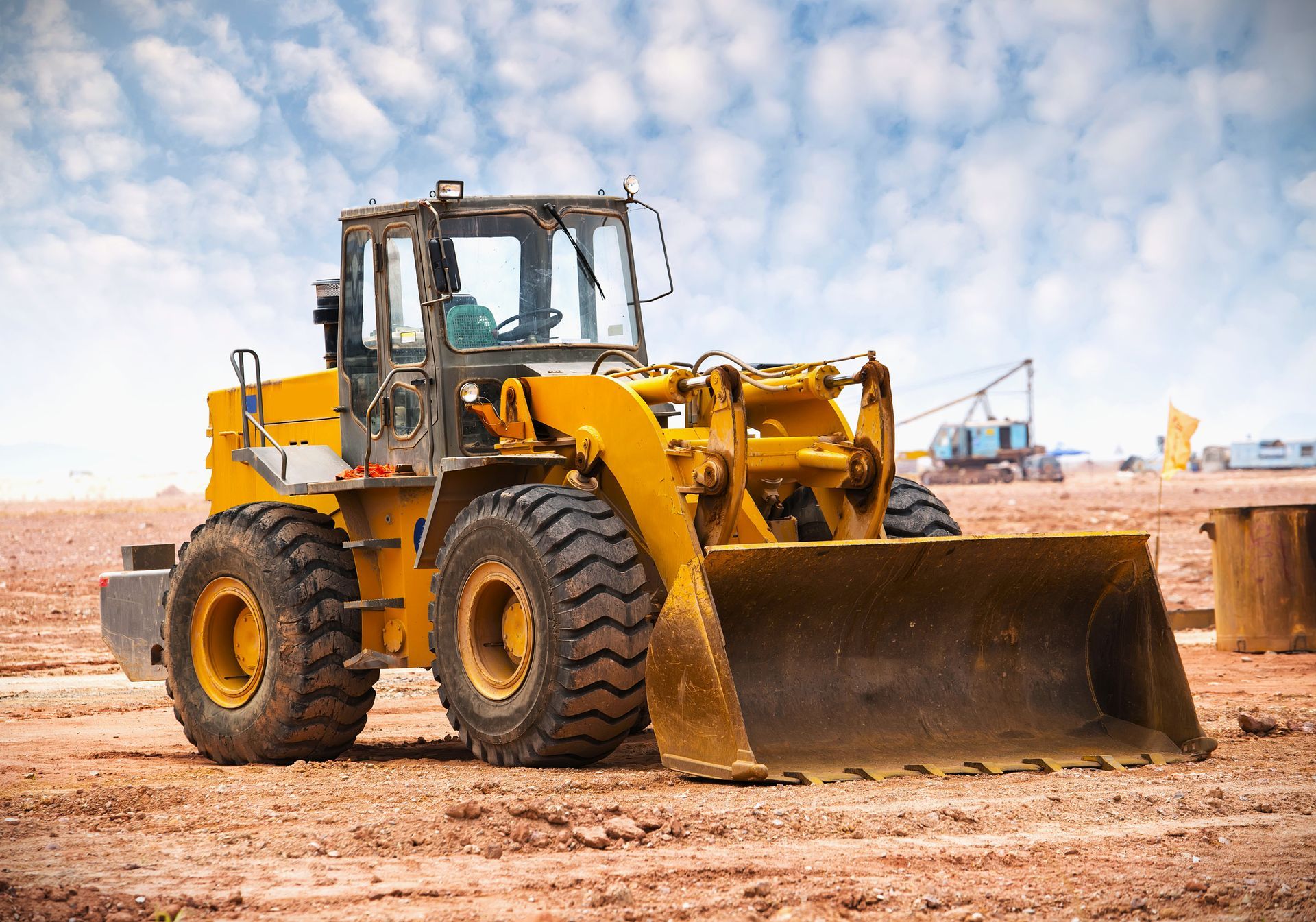 A yellow front-end loader parked on a dirt construction site under a cloudy blue sky.
