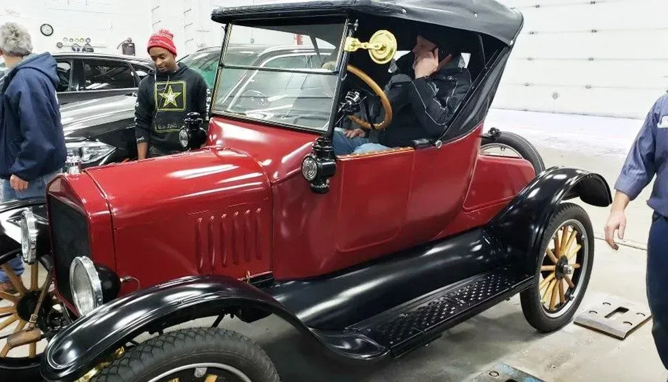 Red and black antique car, with a person seated inside, surrounded by onlookers in a garage.