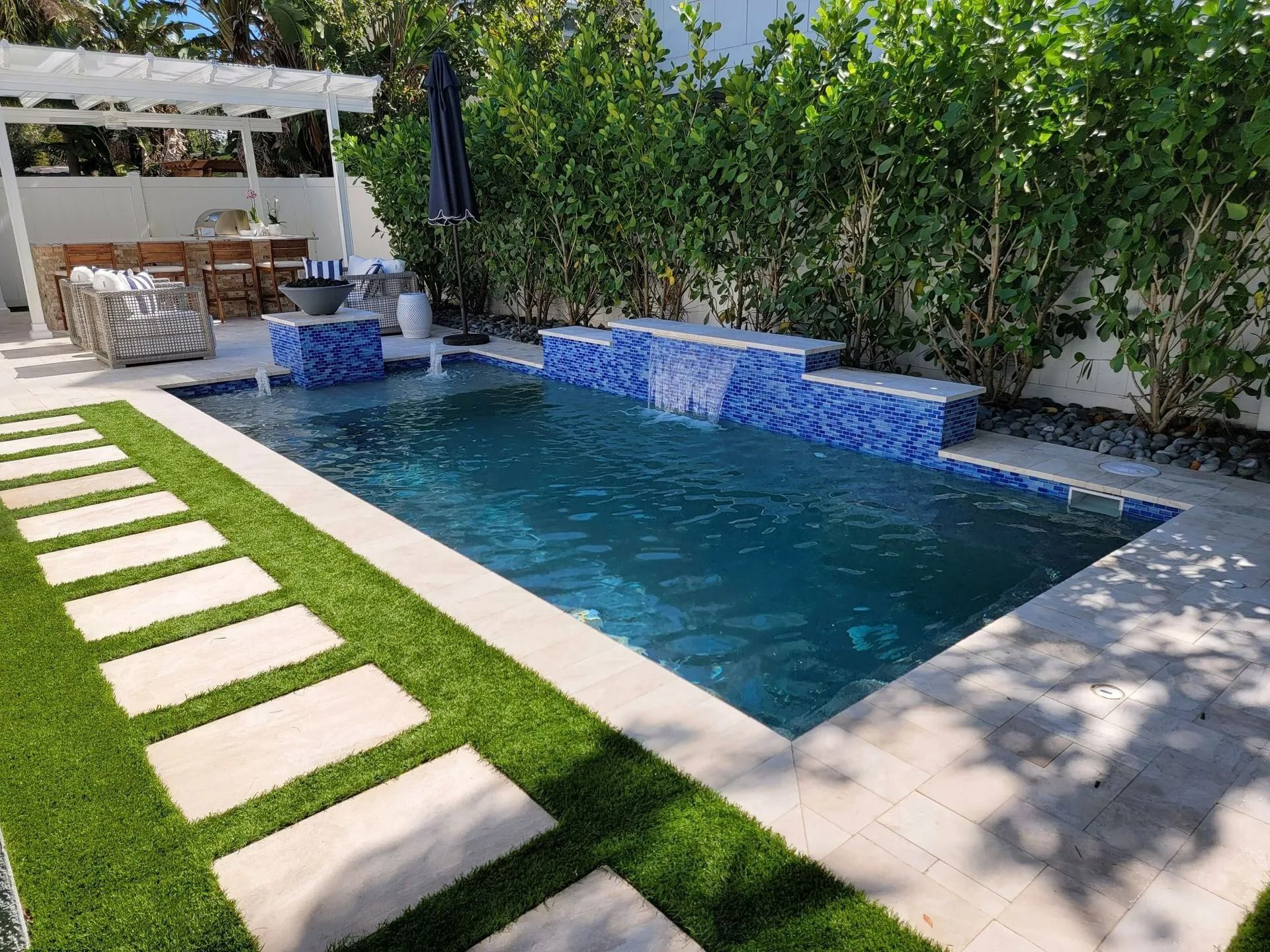 Backyard with pool, pavers, artificial turf, and a waterfall feature; lush greenery surrounds.