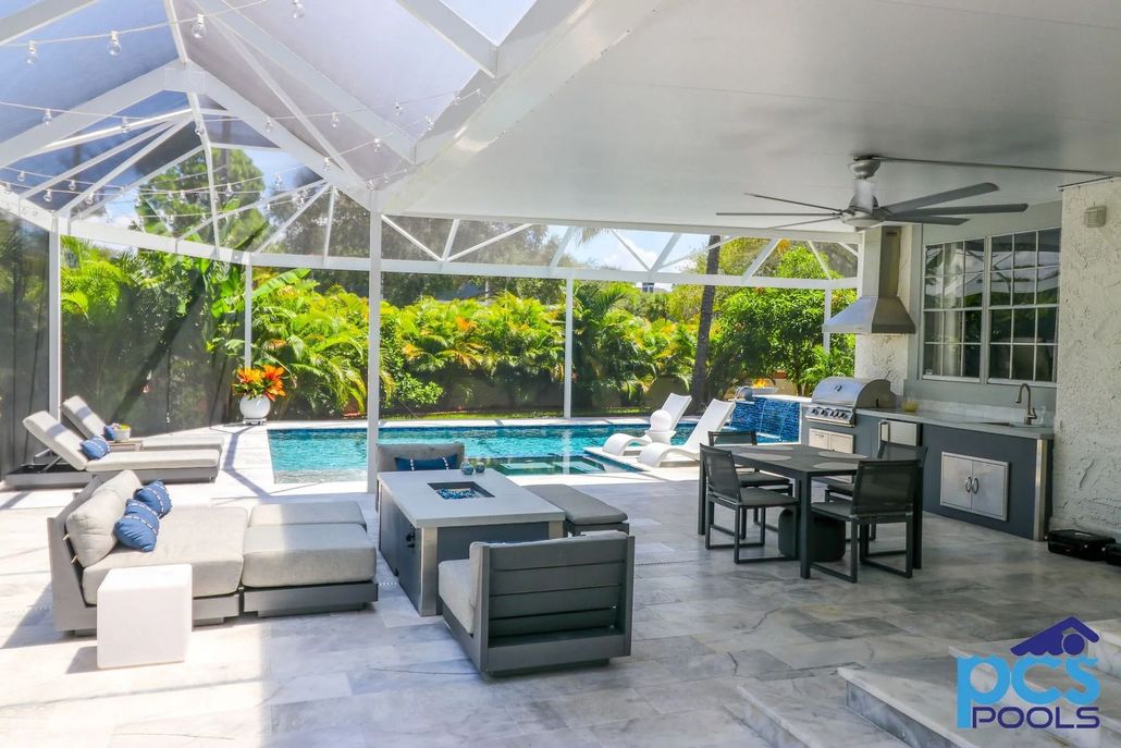 Poolside patio with pool, outdoor furniture, and kitchen. Screened-in enclosure with lush green foliage.