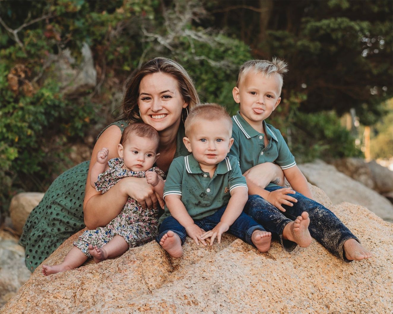 Woman and three young children seated on a large rock; outdoors. The woman smiles, holding a baby. Two boys wear green shirts.