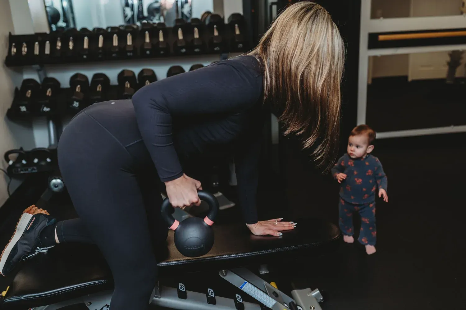 Woman in black workout clothes exercises with a kettlebell in a gym, baby nearby.