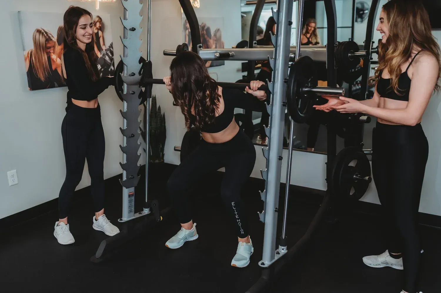 Three people exercising with a weight bar in a gym. One is squatting, while the other two assist and spot.