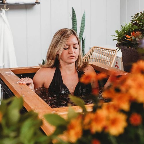 Woman in black swimsuit in wooden tub, eyes closed, surrounded by flowers.