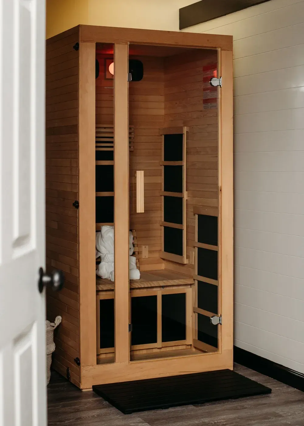 Wooden infrared sauna, open door. Interior view with shelves, towels, and black mat.