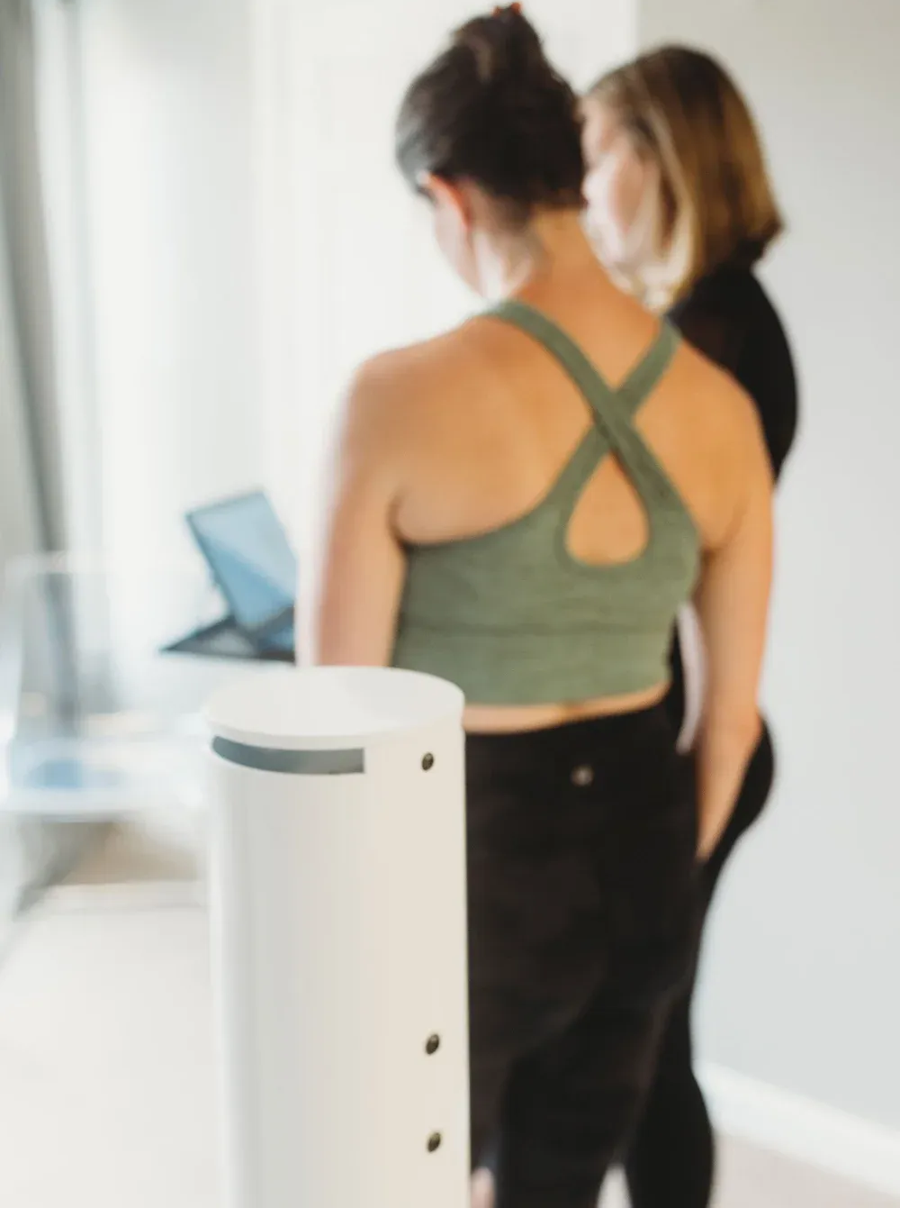 Two people looking at a laptop, near a white cylindrical device. One in green crop top and leggings.