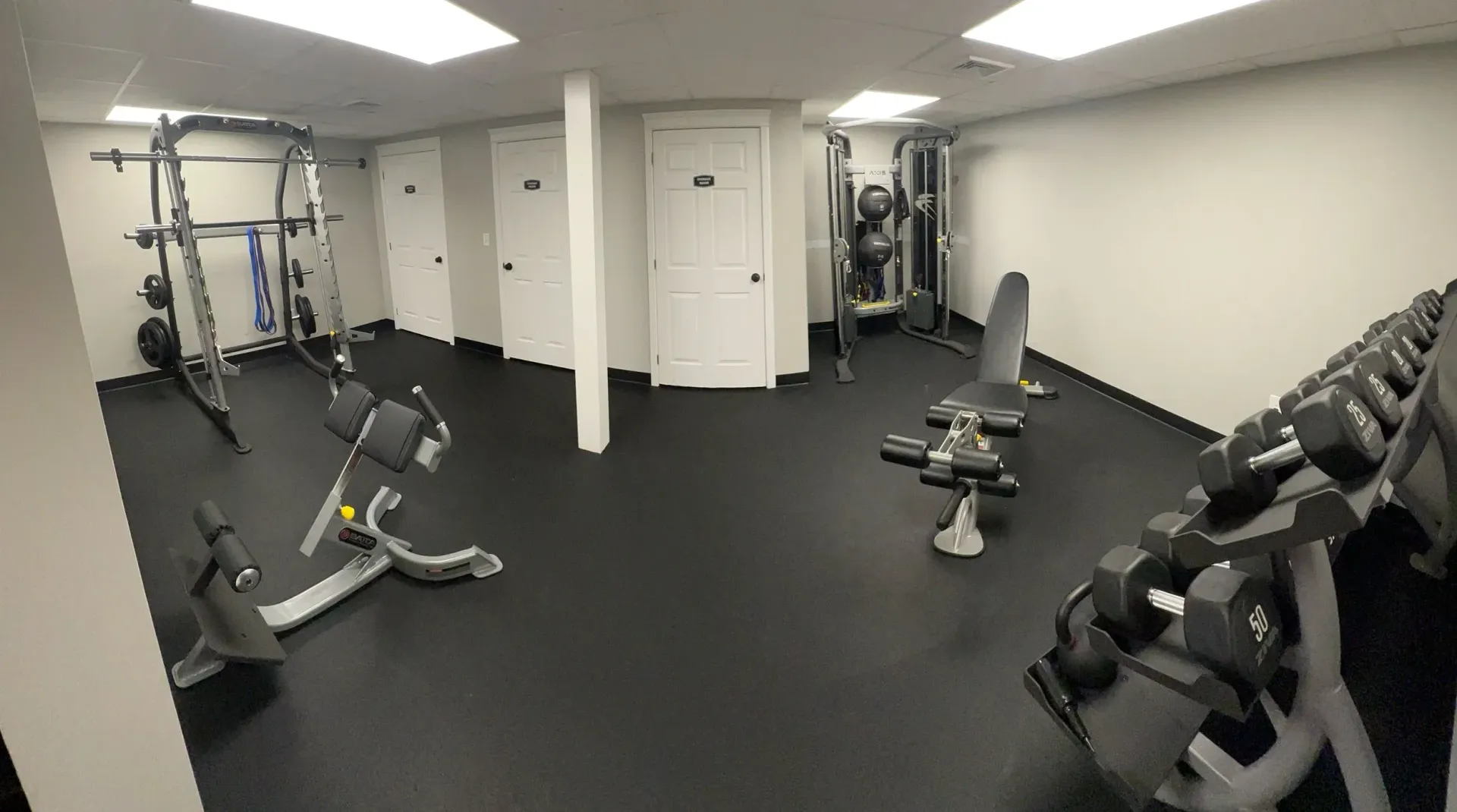 Six women in a gym, selecting dumbbells. They wear black workout clothes, smiling. Gray walls.
