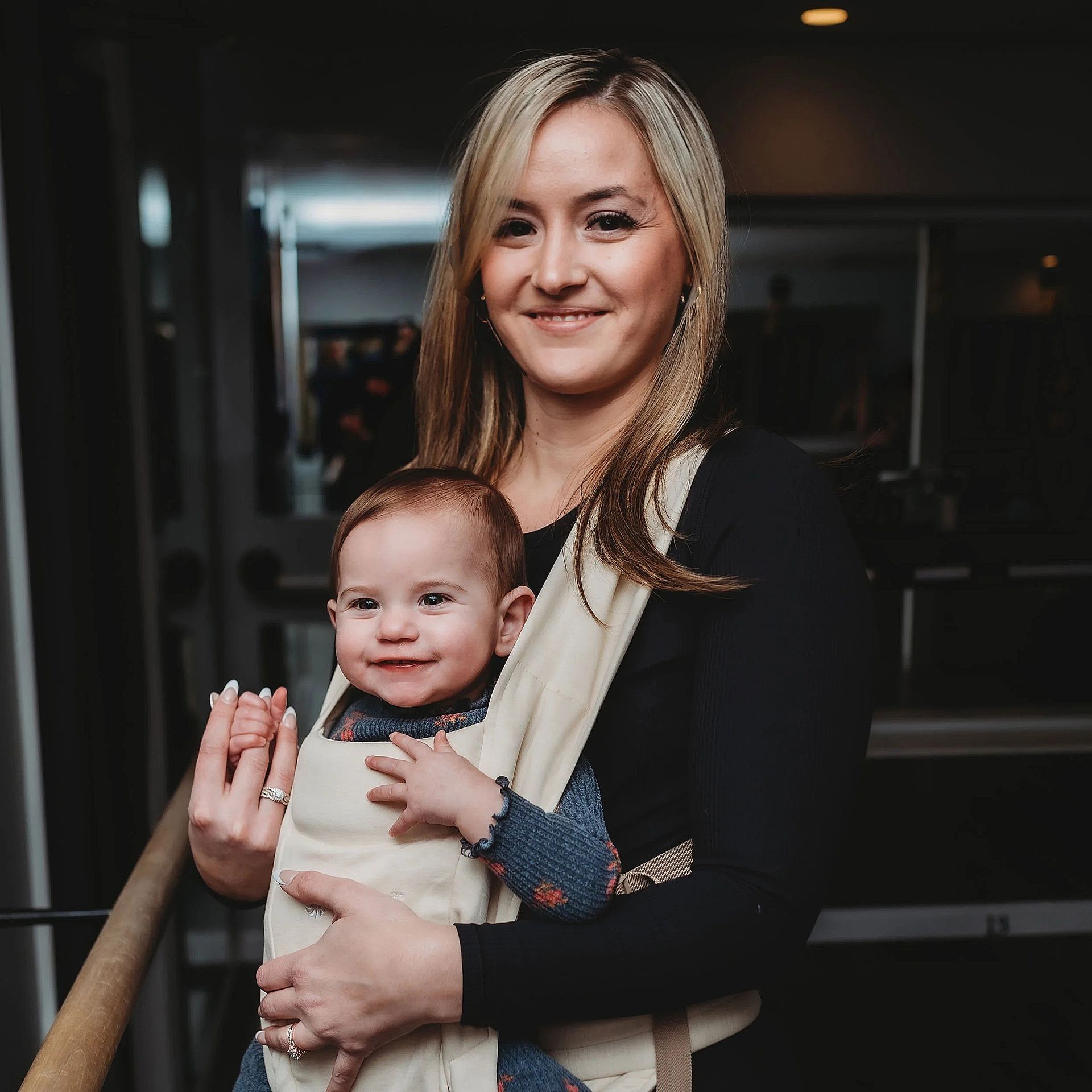 Woman smiling, holding a baby in a beige carrier. Indoors, baby is smiling, woman has blonde hair, wearing a black shirt.