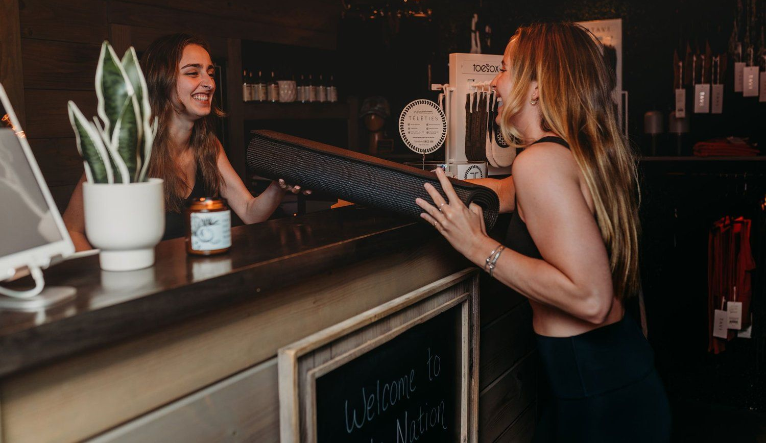 Woman hands yoga mat to another woman at a counter. Store setting with greenery and candle.
