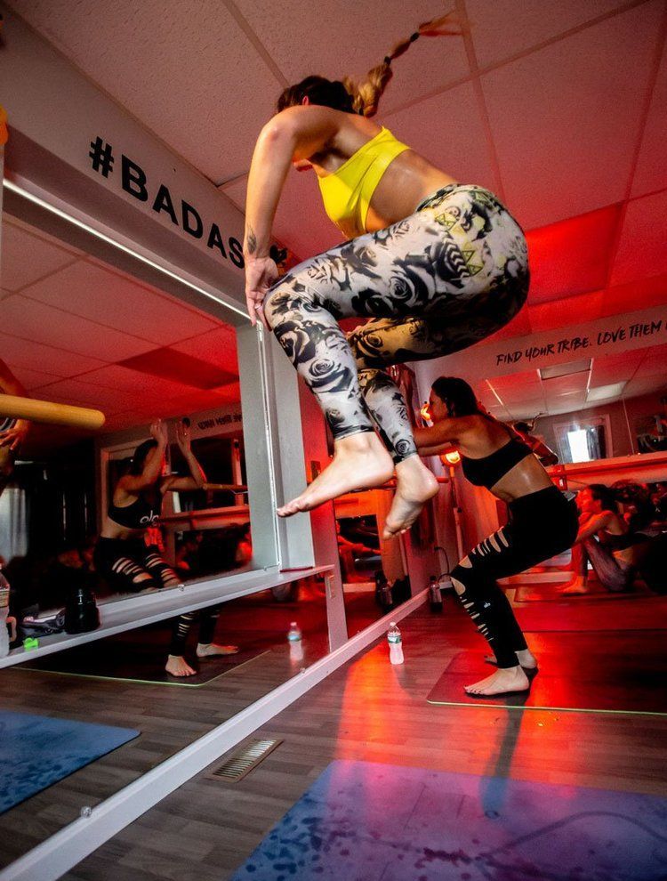 Woman jumps over a barre in a studio, wearing yellow top and patterned leggings. Another person squats nearby.