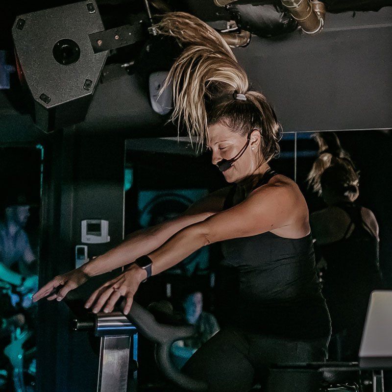 Woman leading a spin class, wearing a headset mic, riding a stationary bike, dimly lit room.