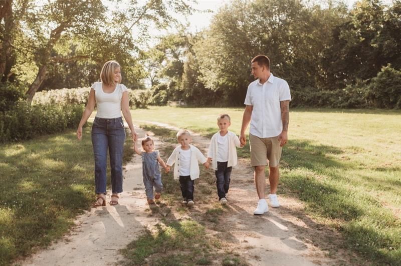 Family of five walking on a dirt path in a sunny park; parents holding hands with three young children.
