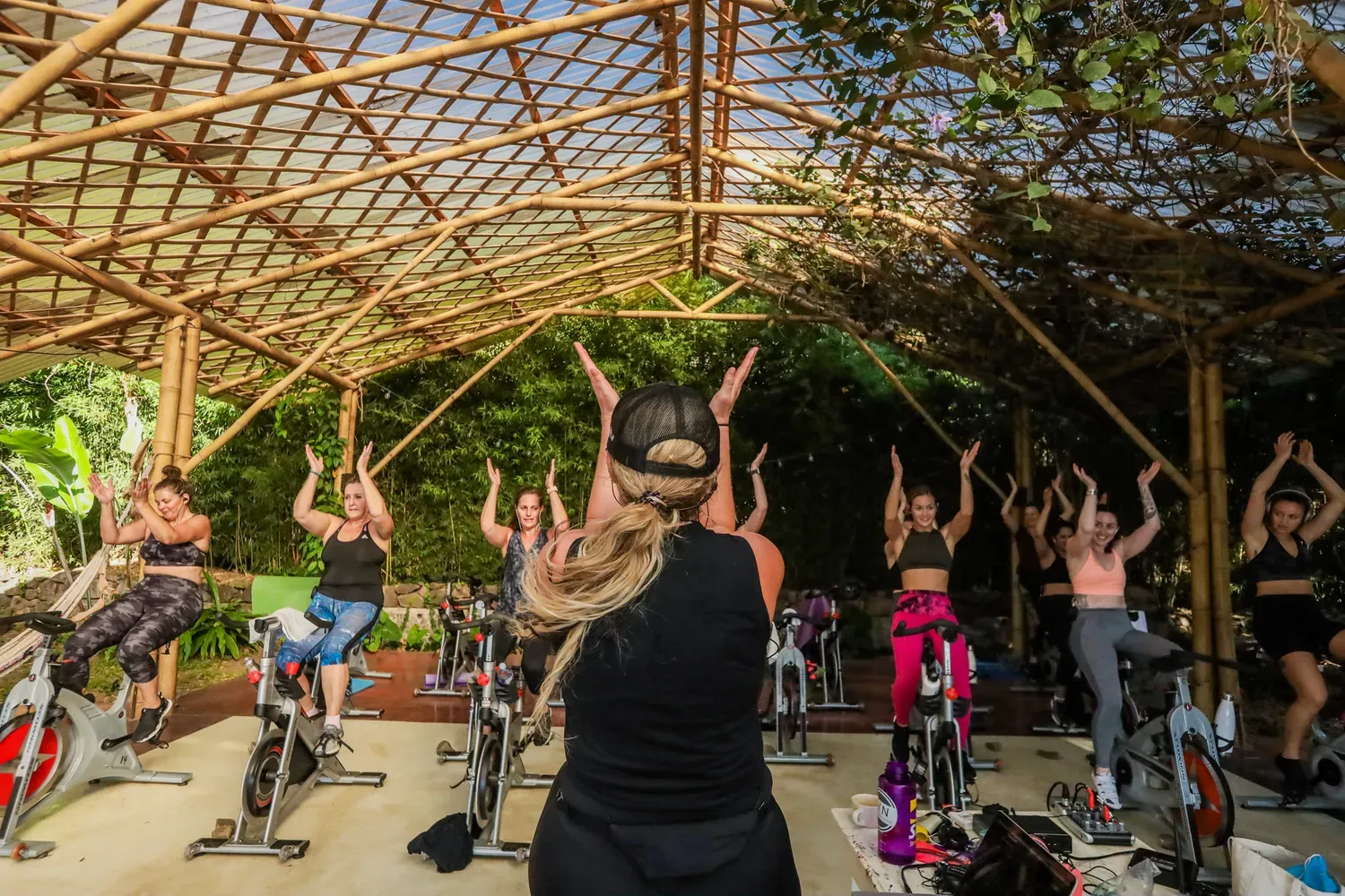 Spin class outdoors under a bamboo structure. Instructor leading class. Participants on bikes with arms raised.