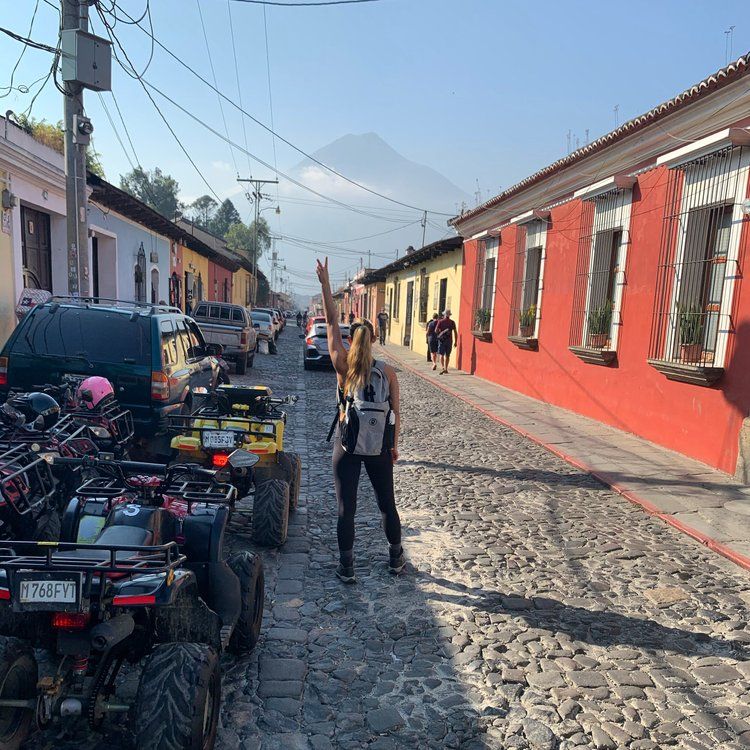 Woman raises arm on cobblestone street, volcano in background. ATV's and cars parked, colorful buildings.