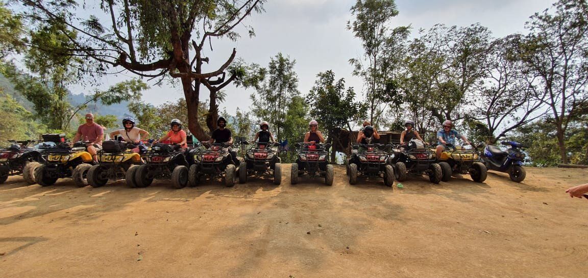 Group of people on ATVs in a dirt area with trees in the background.