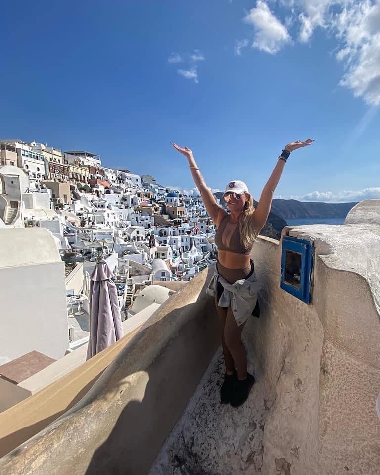 Woman with arms raised, smiles, overlooking whitewashed buildings on Santorini, Greece. Blue sky and sea.