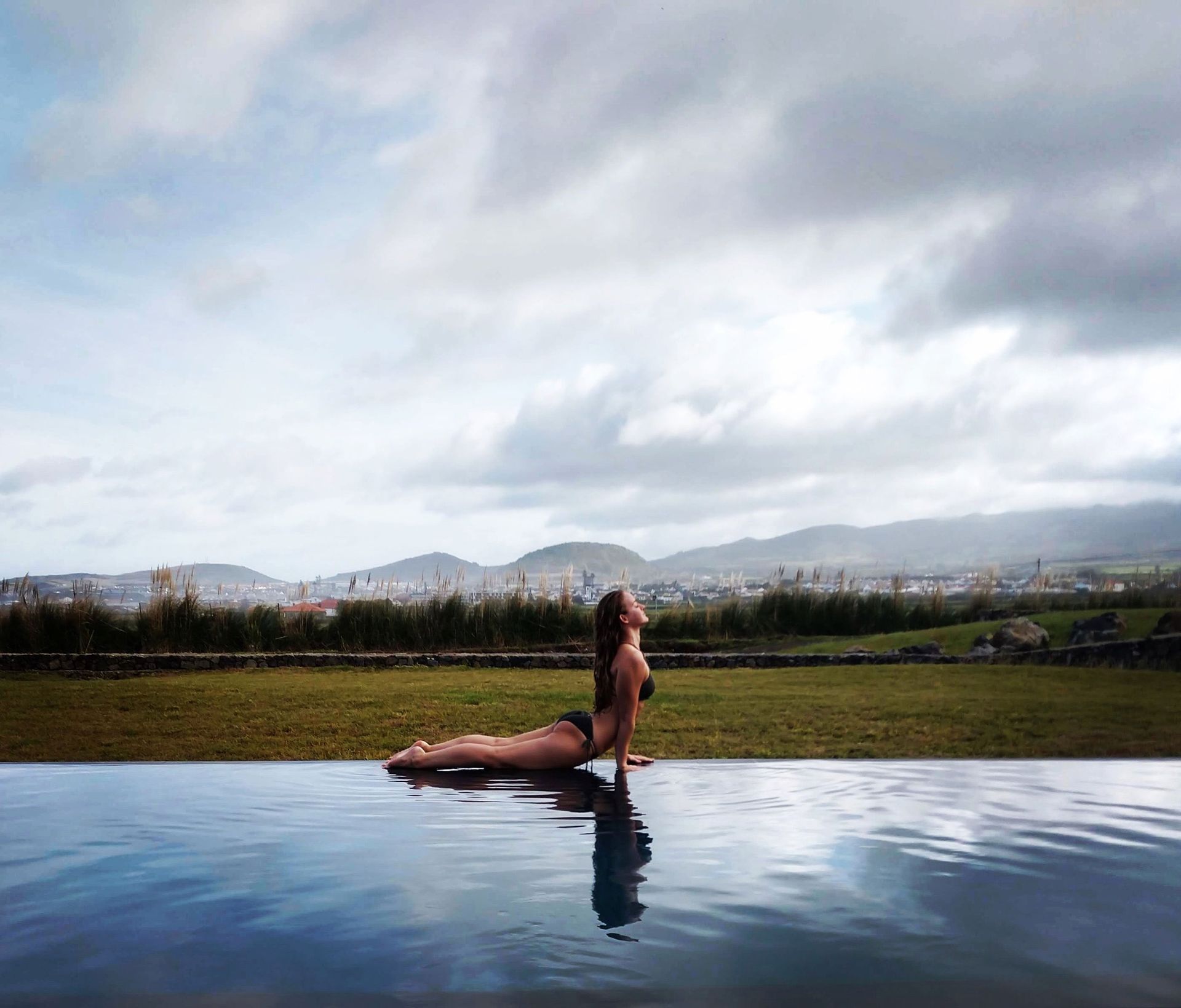 Woman in a black swimsuit does a yoga pose in a pool, mountains in the background, cloudy sky.