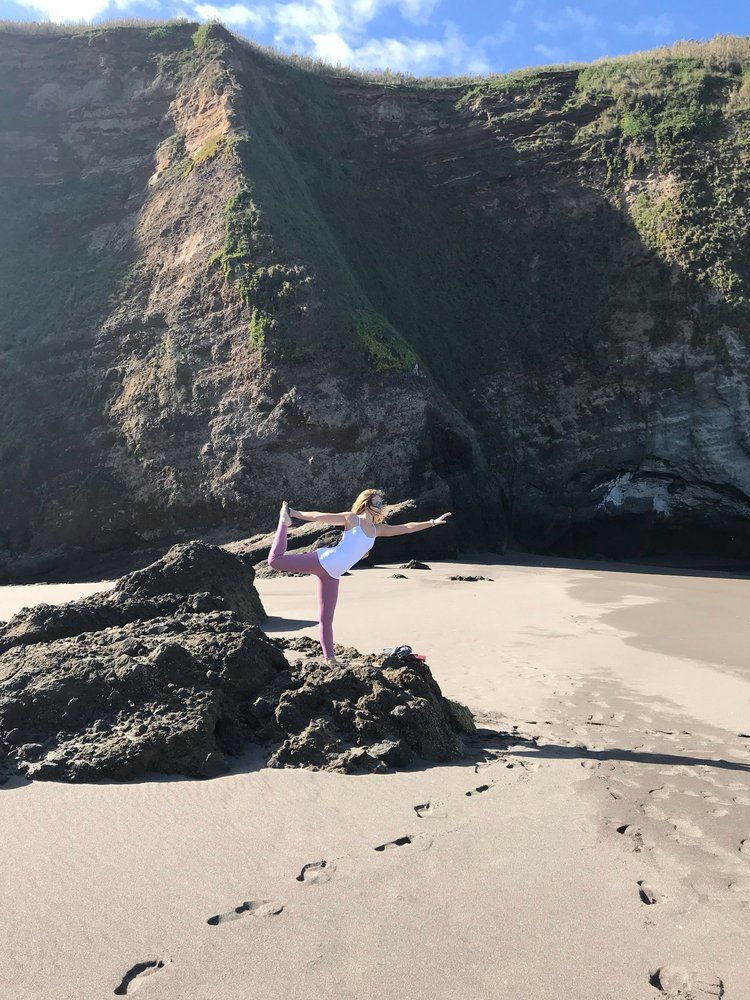 Woman in yoga pose on rock at beach, cliff backdrop.