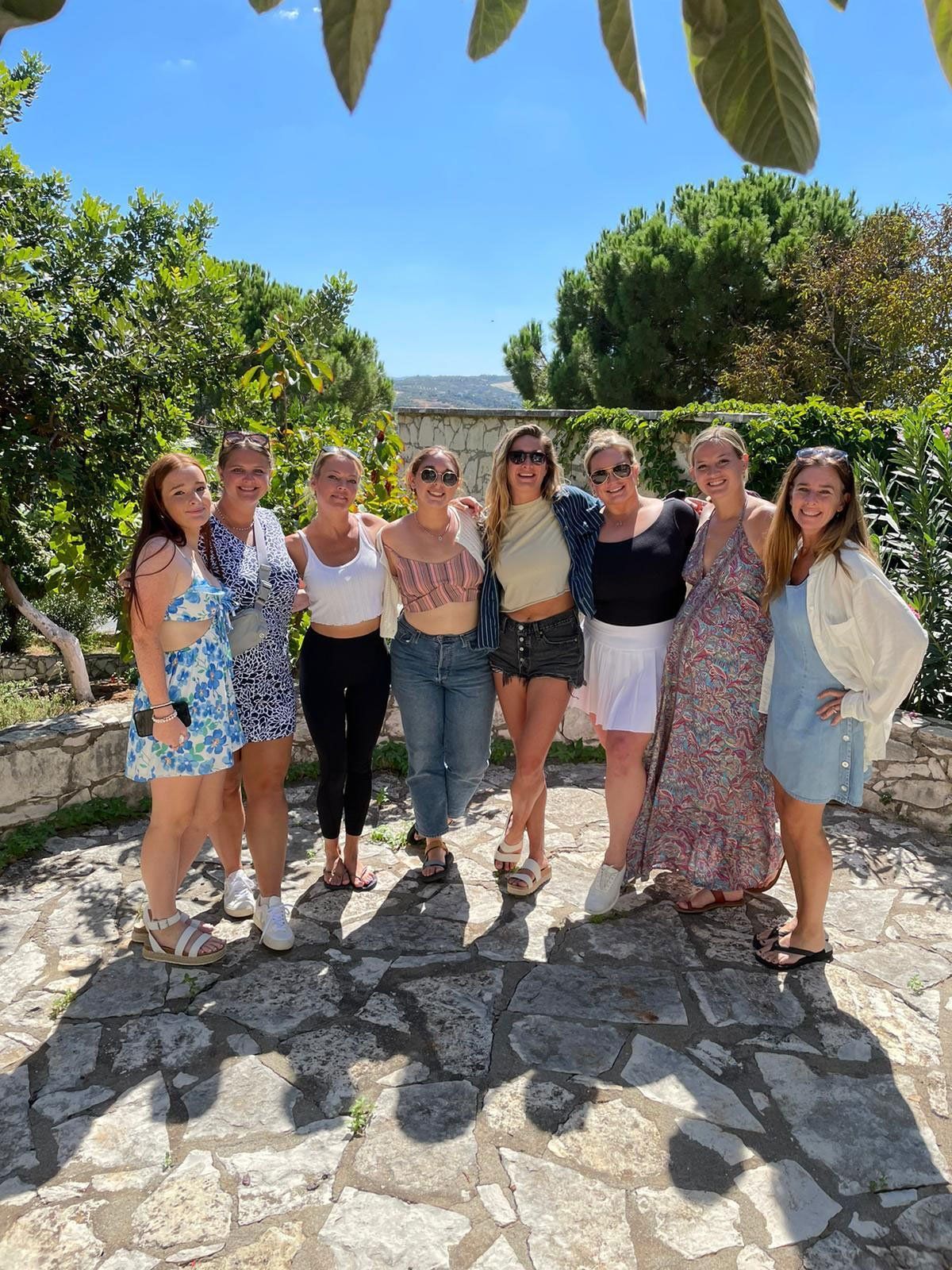 Eight women smiling, posing for a photo outdoors on a sunny day.