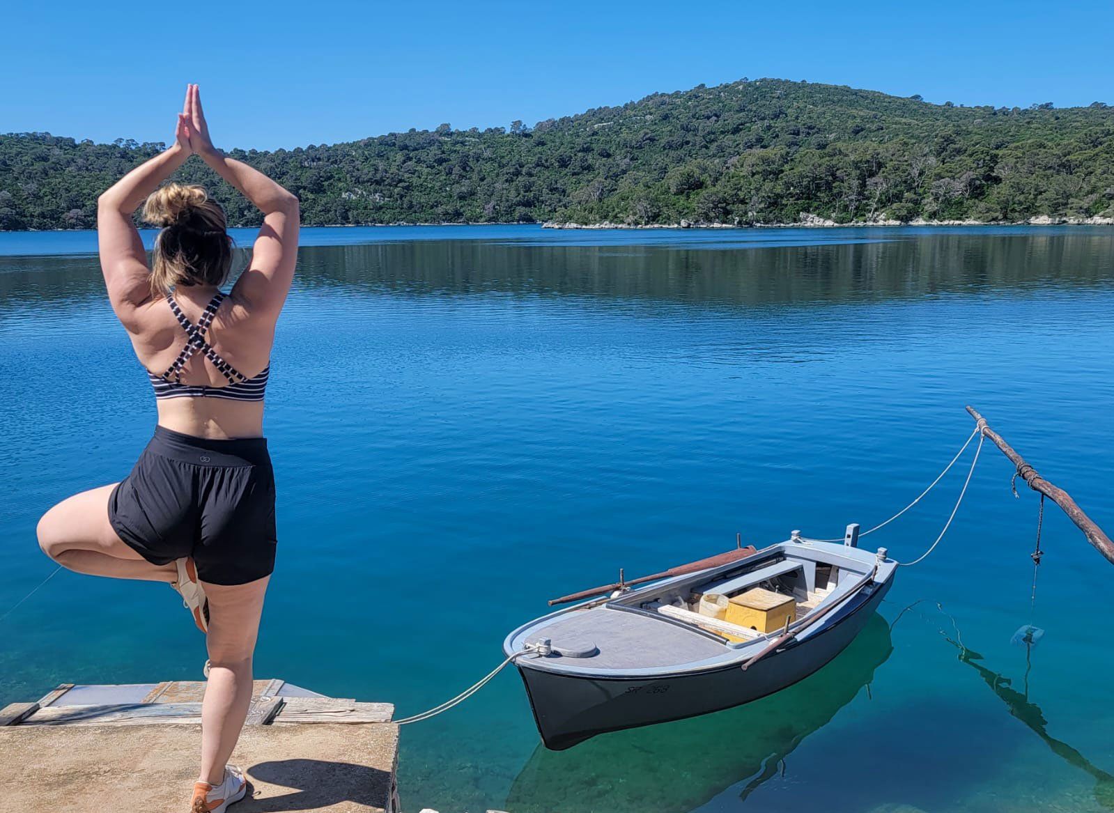 Woman in yoga pose on dock, with boat on blue water, forested hills in background.