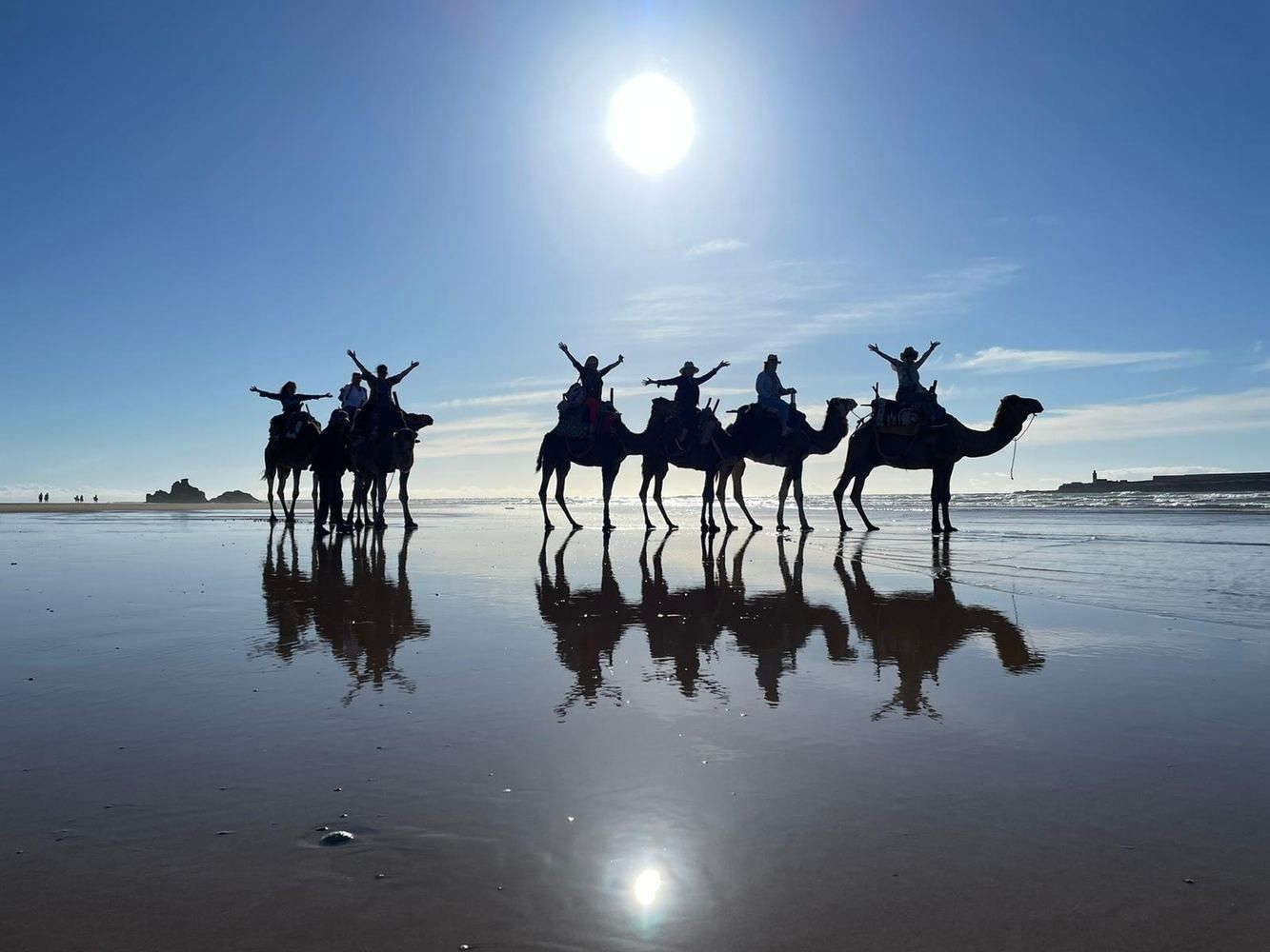 People riding camels on a beach, silhouetted against a bright sun and sky, reflecting in the wet sand.