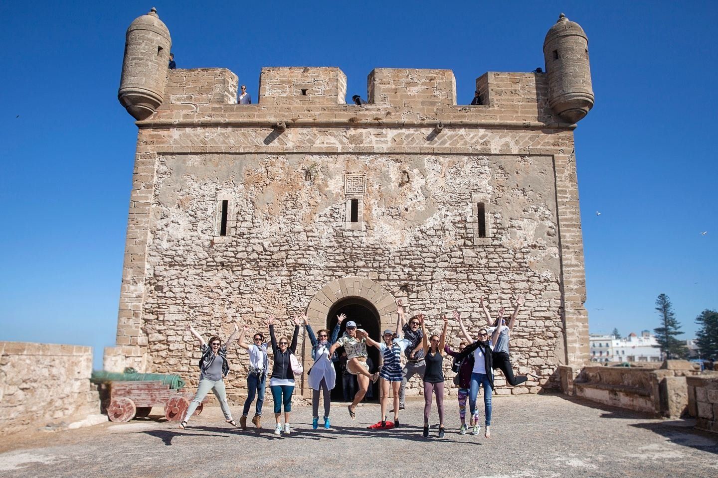 Group of people jumping in front of a stone fortress with two towers; sunny, blue sky.