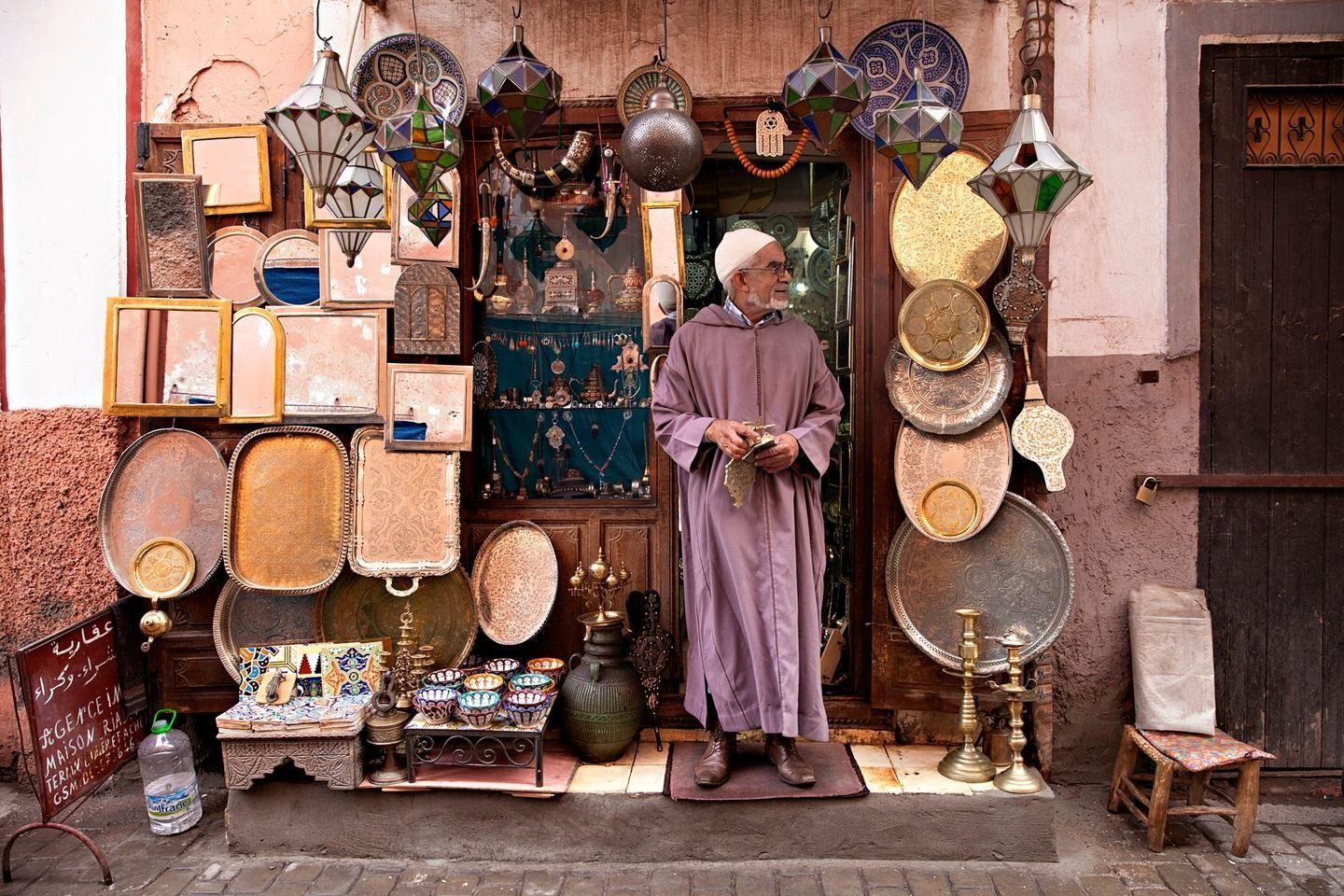 Man in purple robe stands in doorway of shop in Morocco, surrounded by metalwork and lanterns.