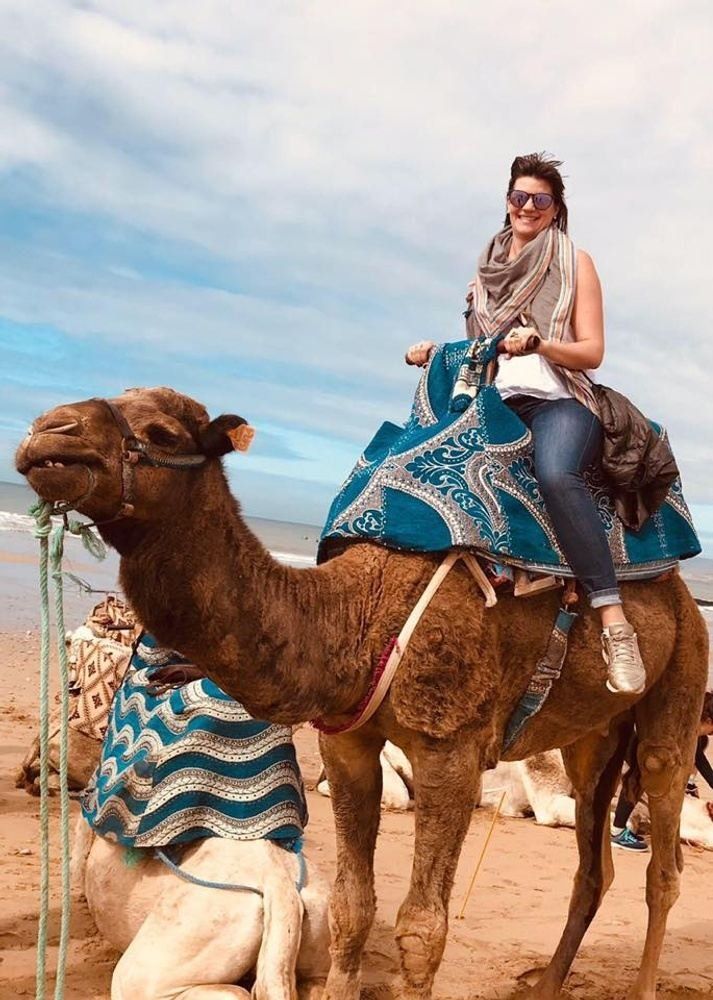 Woman riding a camel on a beach, smiling. Blue and white blanket on camel.