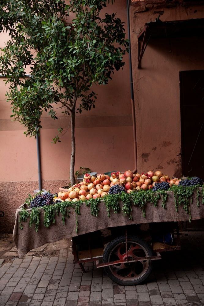 Fruit cart with pomegranates and grapes under a tree, set on a brick street with pink walls.
