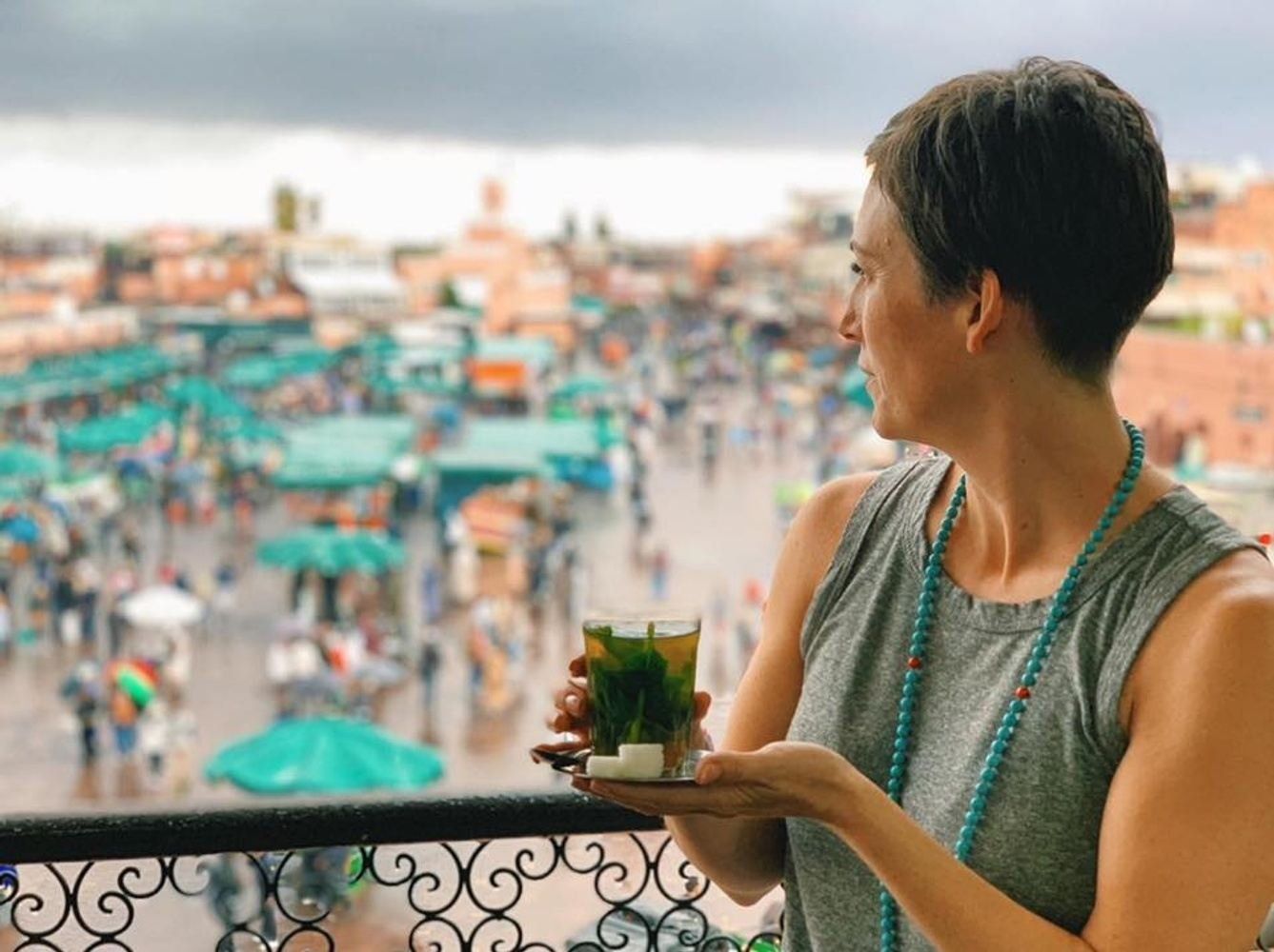 Woman on balcony holding tea, overlooking crowded marketplace, turquoise jewelry.