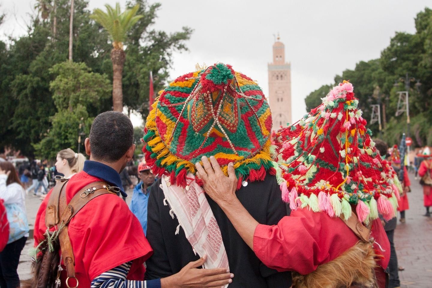 Men wearing colorful hats and red outfits in a crowded outdoor setting, with a tall minaret in the background.