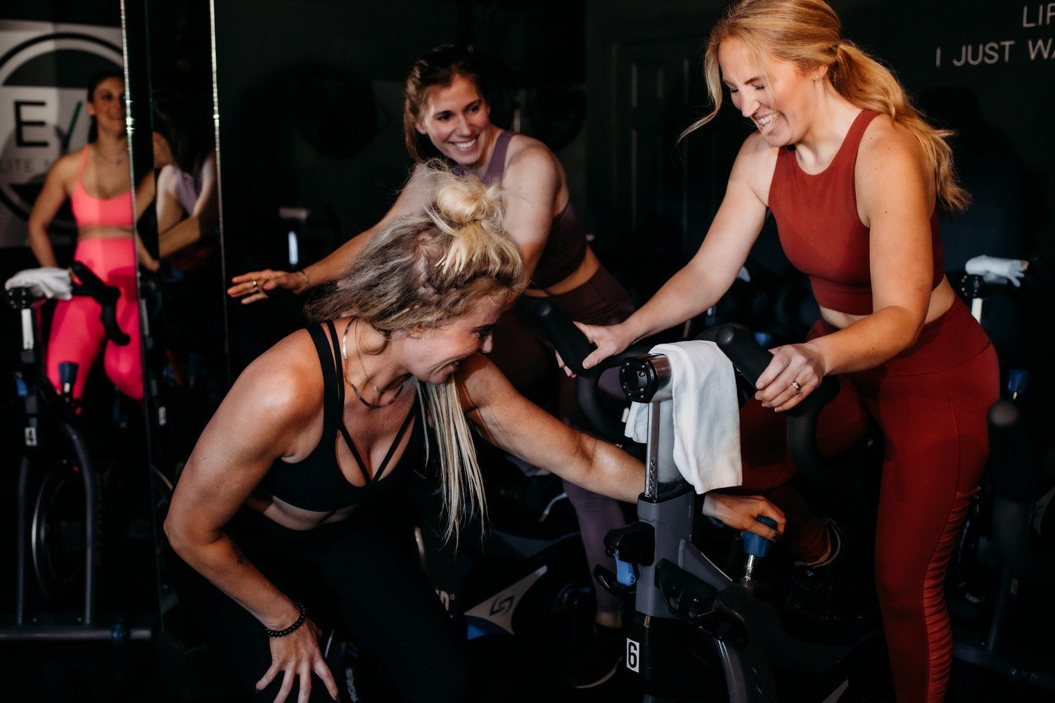 Women in activewear laughing and smiling during an indoor cycling class.