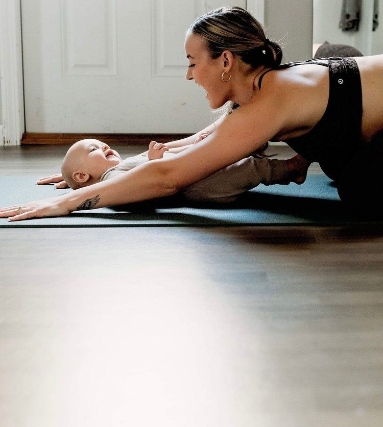 Woman and baby on yoga mat, arms extended, laughing in sunlit room.