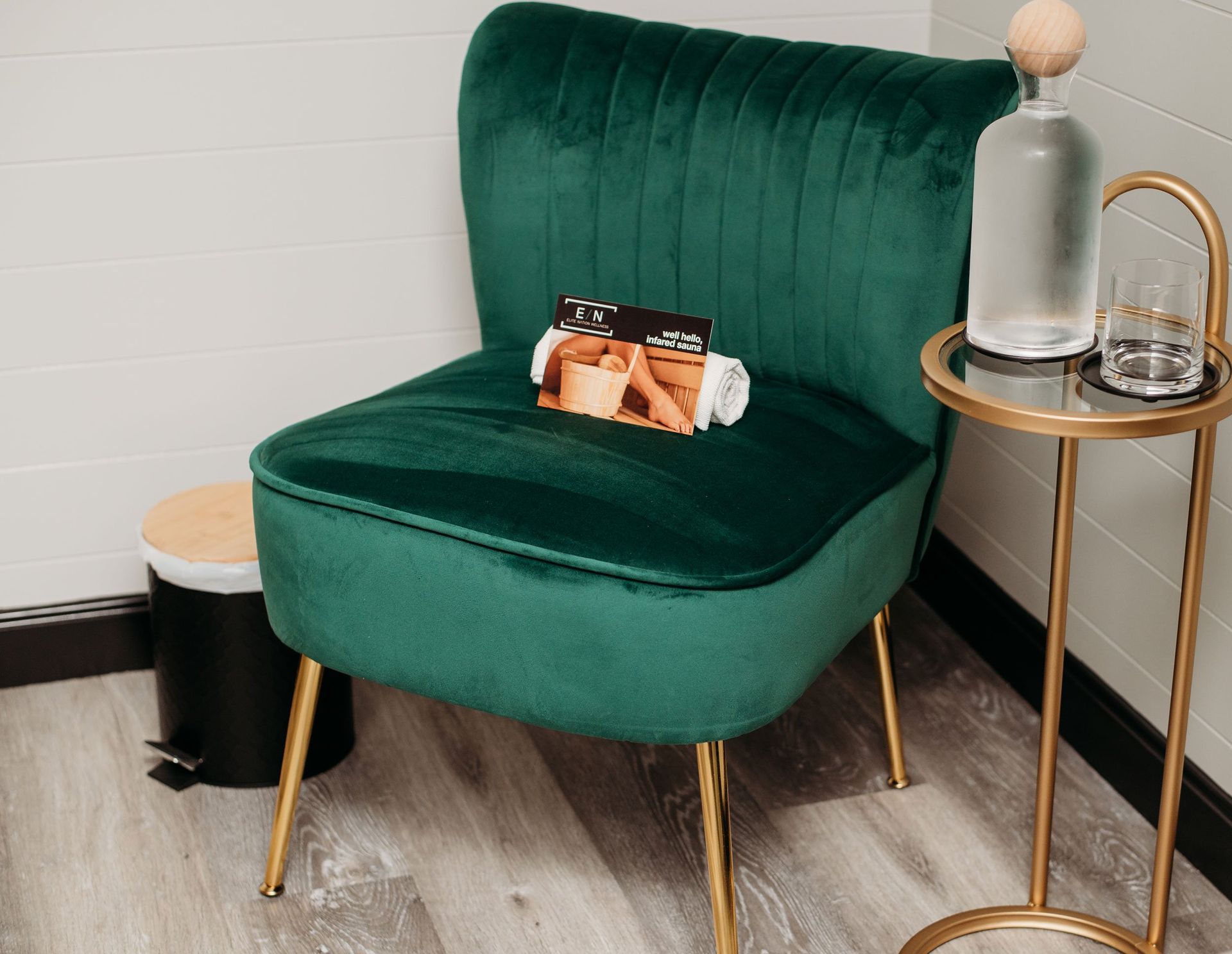 Emerald green velvet chair with gold legs, small gold side table, bottle, and glass in a corner.