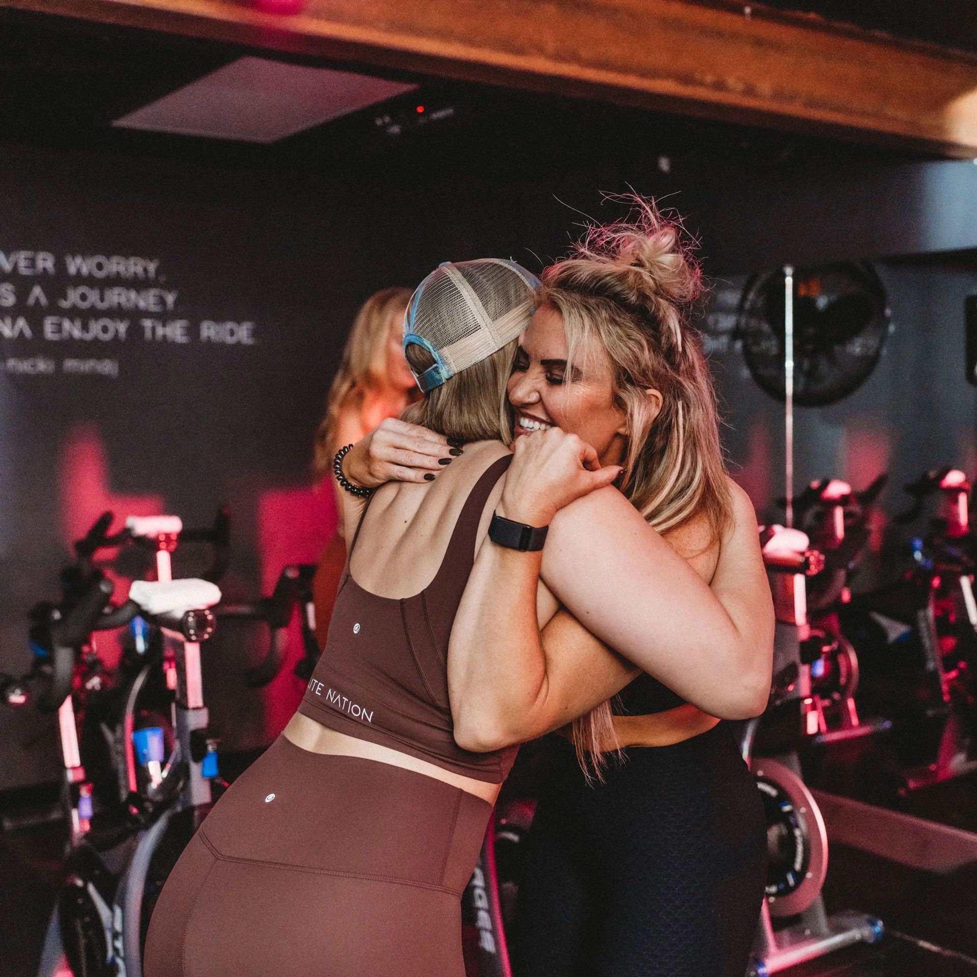 Two women embracing in a spin class studio. One wears brown workout attire, the other black. Dimly lit with bikes.