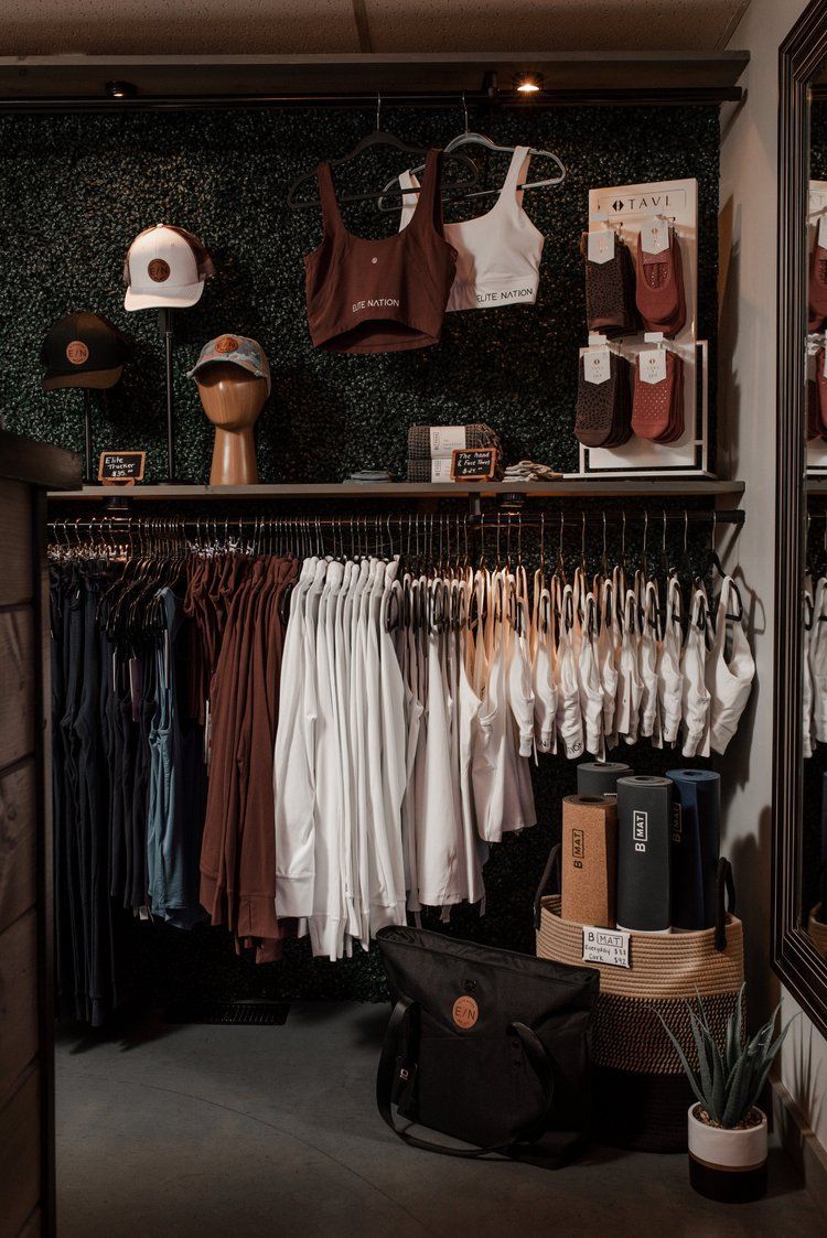 Clothing store display with apparel: shirts, hats, sports bras, and accessories on racks and shelves. Green wall background.