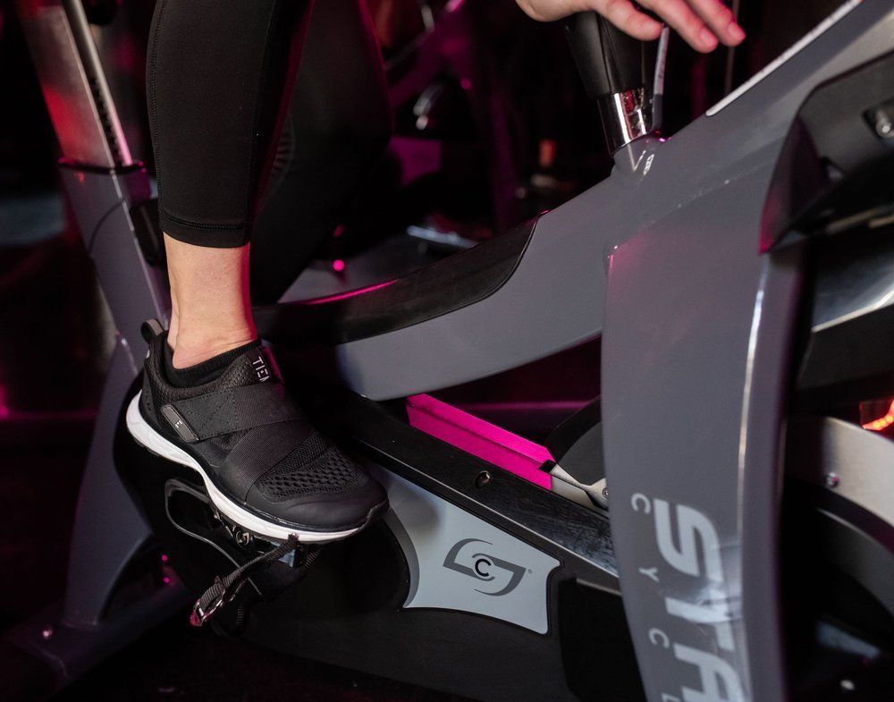 Close-up of a person's foot in a black athletic shoe on a stationary bike pedal, grey machine, indoors.